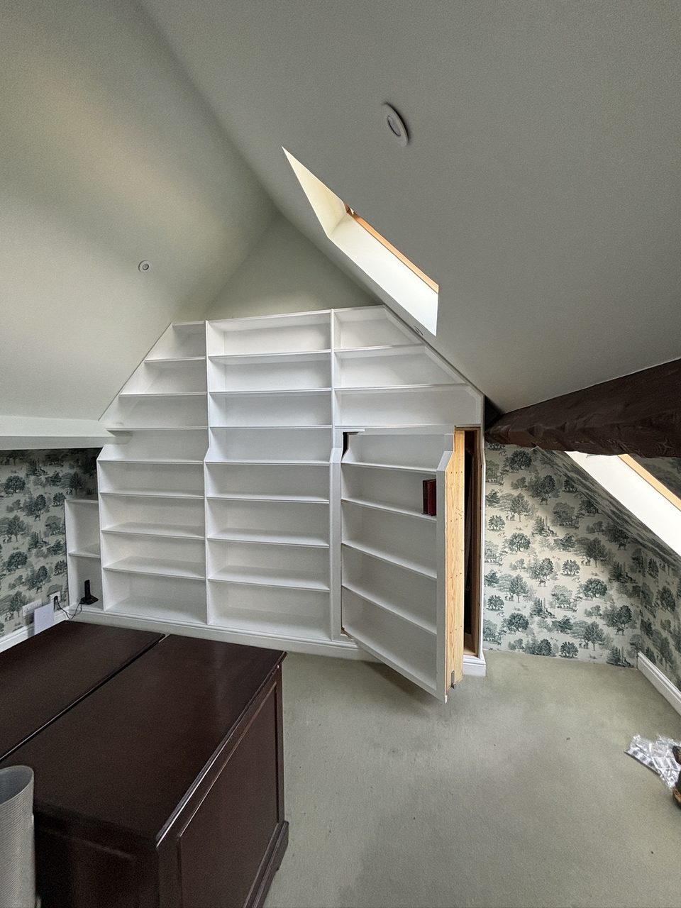 Built-in white bookcase fitted into a loft gable, with one section hinged open as a hidden door