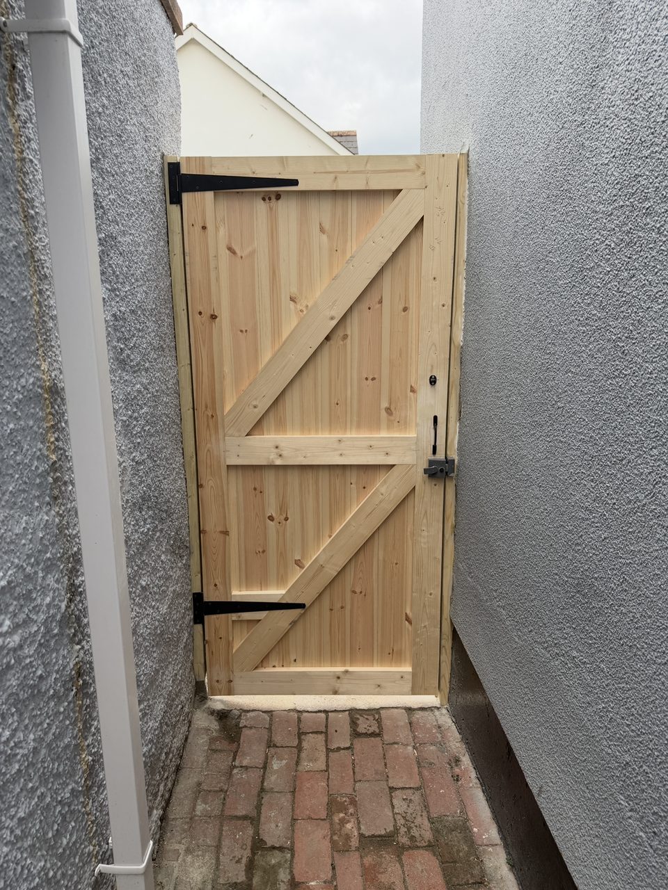 Timber side gate with black T-hinges and a latch, fitted between two walls